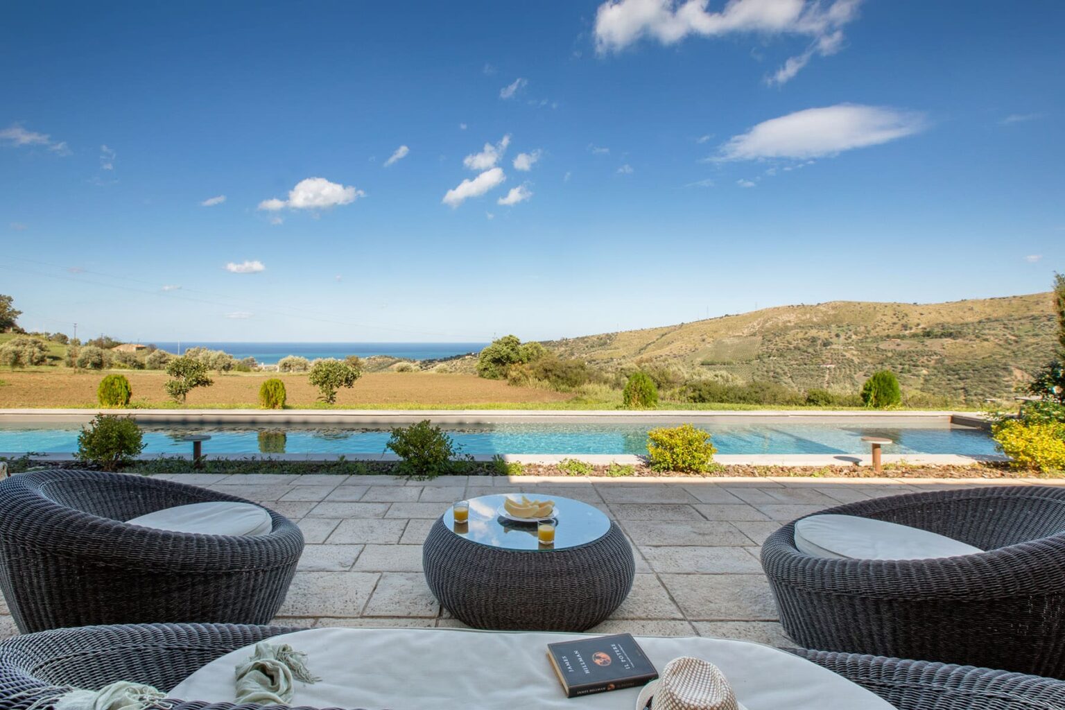 Modern outdoor lounge area with wicker chairs and a coffee table, overlooking a serene pool and scenic landscape with hills and ocean in the background. Ideal for relaxation and enjoying nature. | Only on SopranoVillas