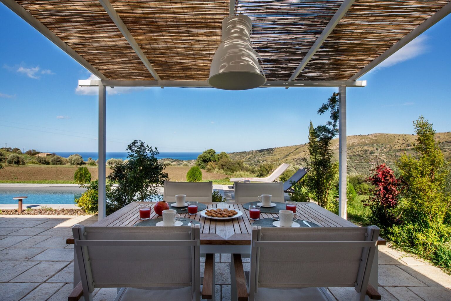 Outdoor dining area with a wooden table set for a meal, featuring refreshing drinks, surrounded by lush greenery and a scenic view of the pool and hills under a clear blue sky. | Only on SopranoVillas