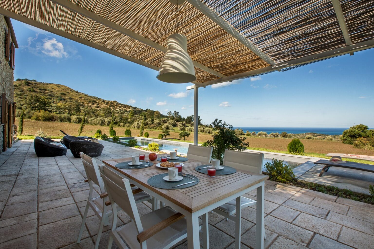 Outdoor dining area with a wooden table set for breakfast, featuring plates and drinks, under a shaded pergola. Scenic view of a lush landscape and ocean in the background, perfect for relaxation and entertaining. | Only on SopranoVillas