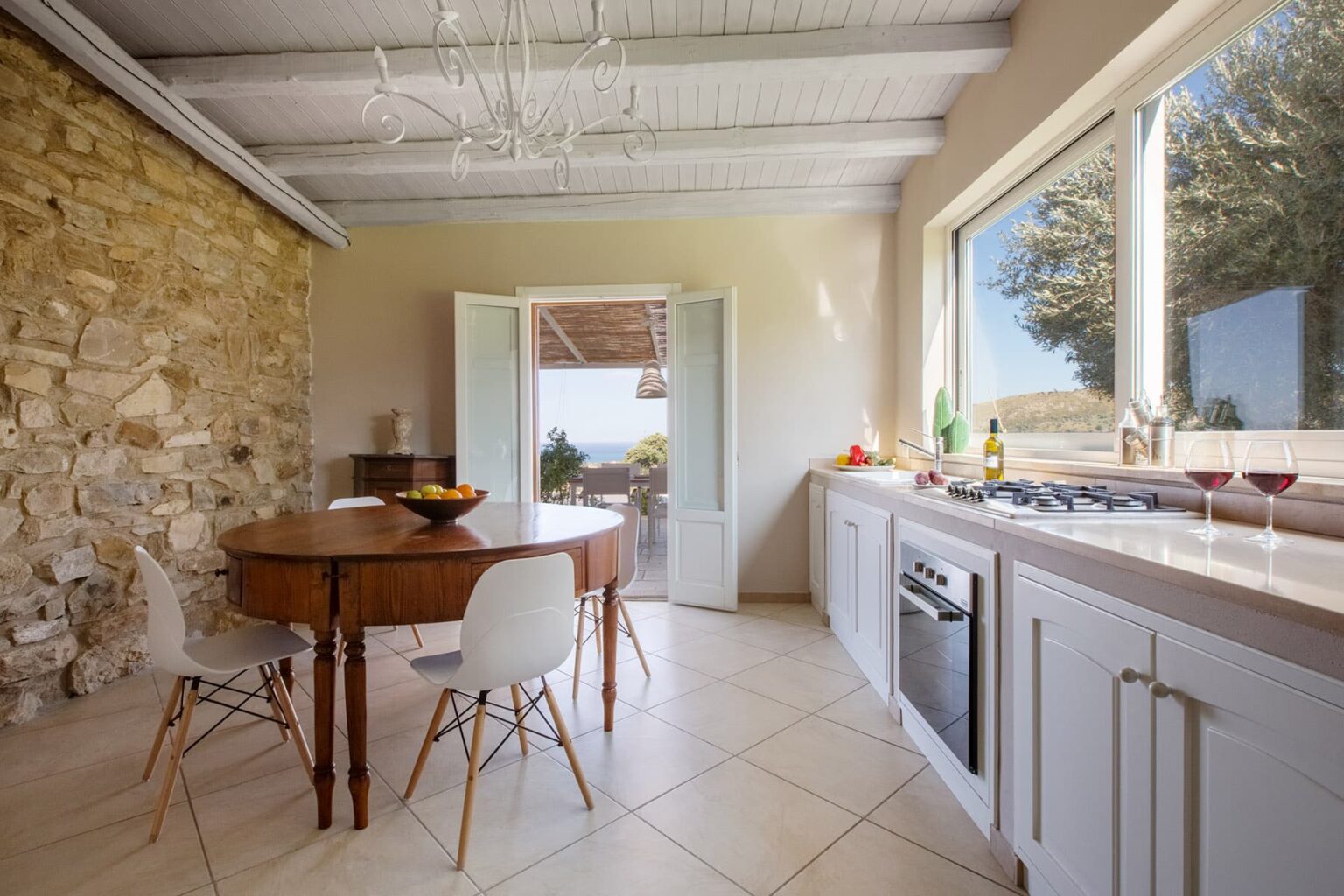 Bright and modern kitchen with a stone wall, featuring a wooden dining table and stylish chairs, large windows offering a scenic view, and contemporary appliances. | Only on SopranoVillas
