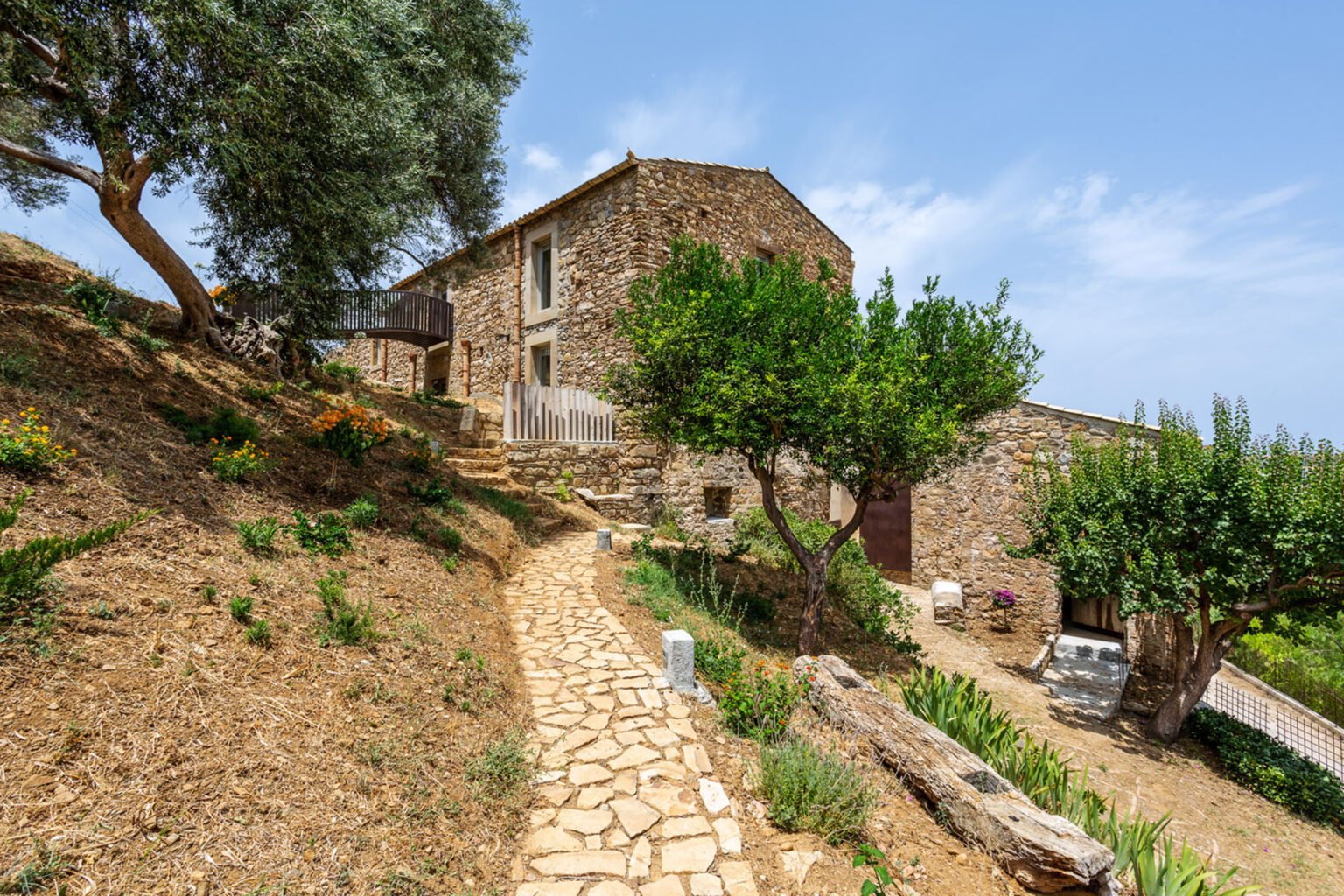 Scenic view of a rustic stone house surrounded by greenery, featuring a winding pathway, olive trees, and a clear blue sky. Ideal for showcasing rural architecture and landscape design. | Only on SopranoVillas