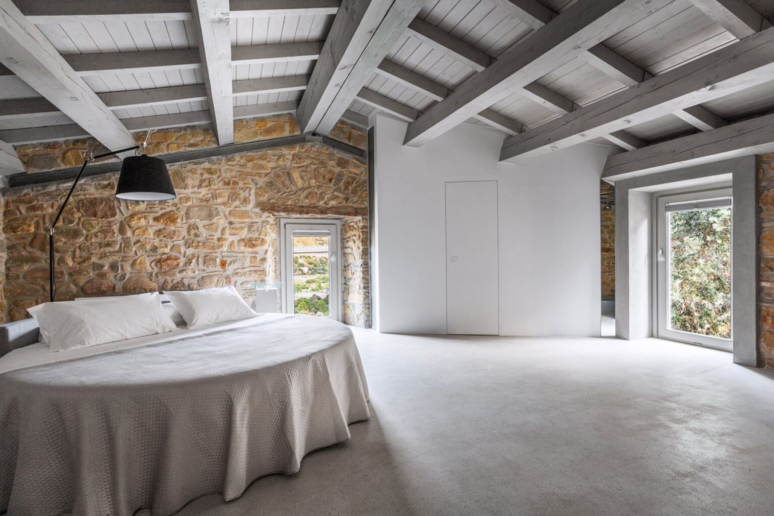 Modern bedroom interior featuring a round bed with a light-colored bedspread, exposed stone walls, and wooden beams. The room has large windows allowing natural light, creating a serene and spacious atmosphere. | Only on SopranoVillas