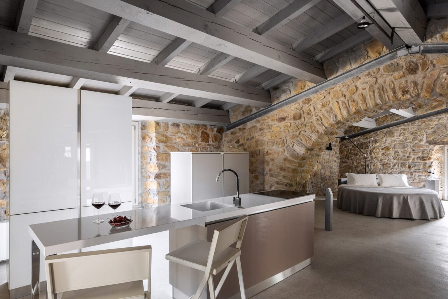 Modern kitchen and bedroom area in a stylish stone-walled apartment, featuring a sleek kitchen island, white refrigerator, and a cozy bed with a rounded design. Ideal for a relaxing getaway. | Only on SopranoVillas