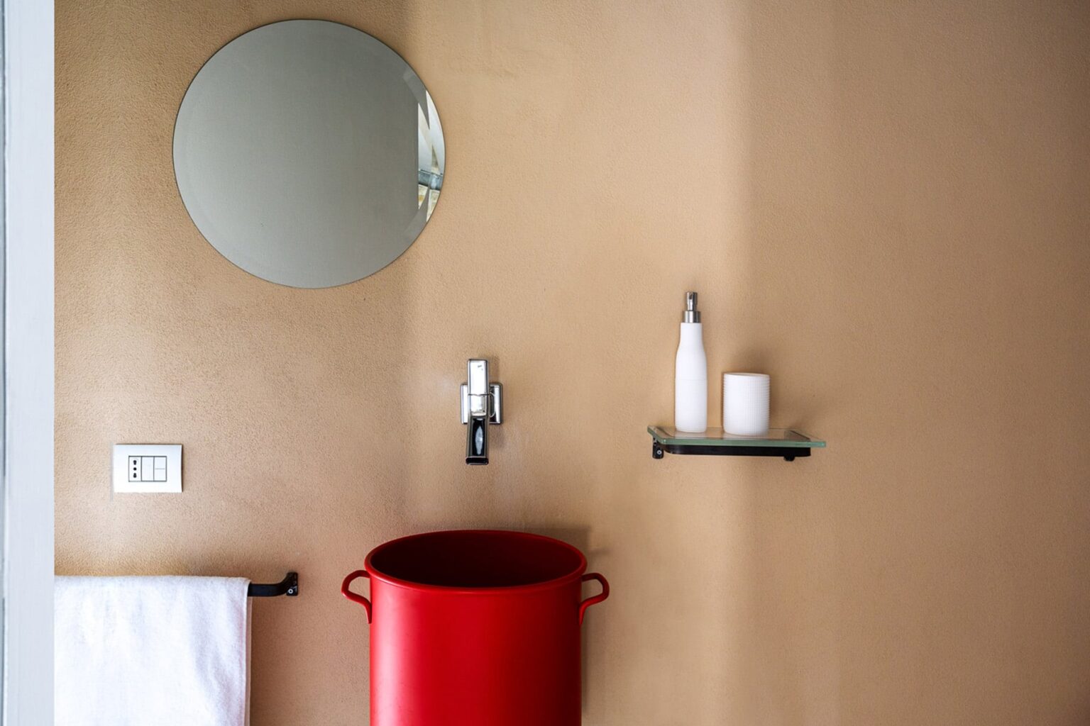 Modern bathroom interior featuring a round mirror, a red bucket, a white towel, and a minimalist shelf with toiletries against a beige wall. | Only on SopranoVillas