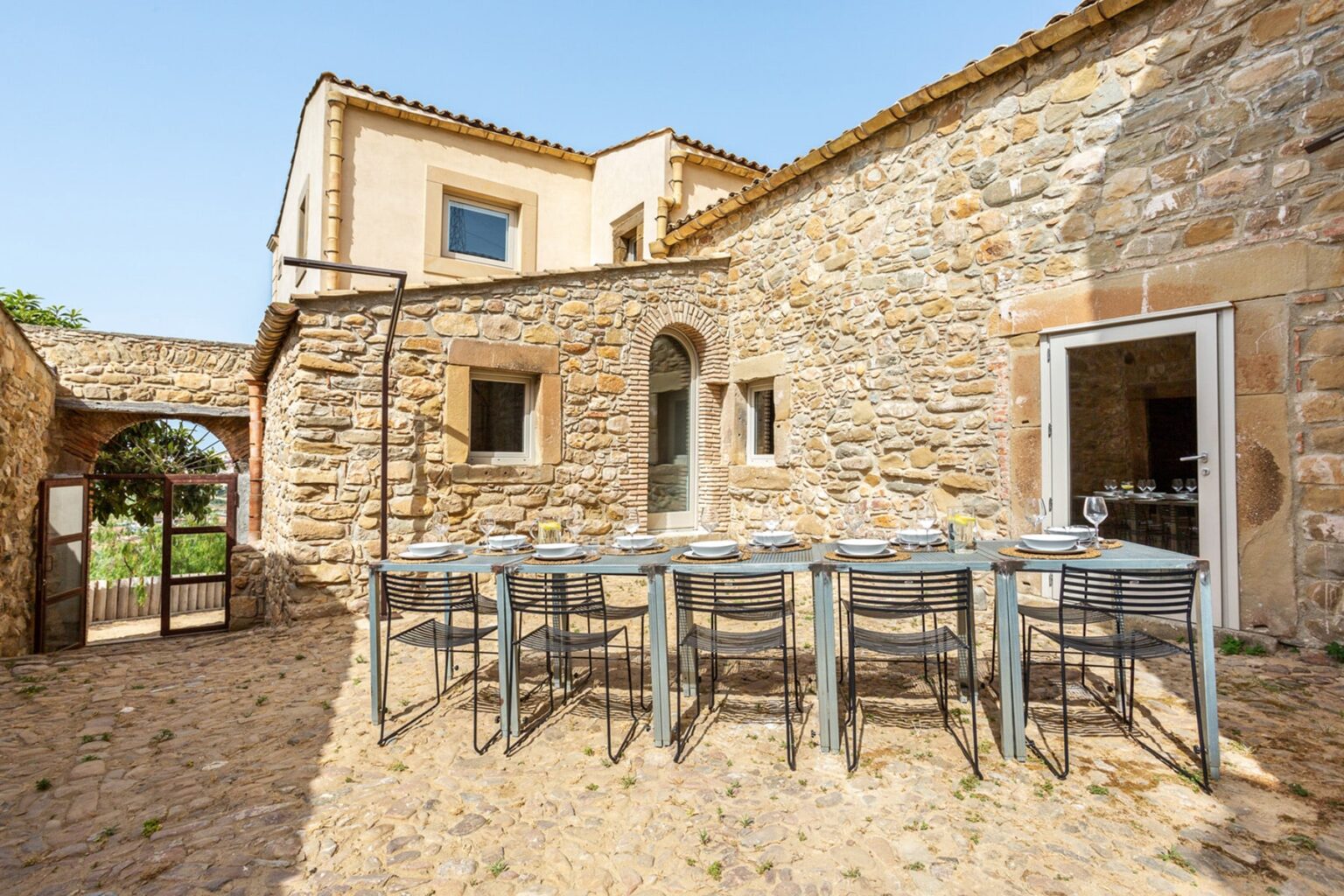 Outdoor dining area with a long metal table set for a meal in front of a rustic stone house, surrounded by greenery and blue skies. Ideal for gatherings and al fresco dining. | Only on SopranoVillas