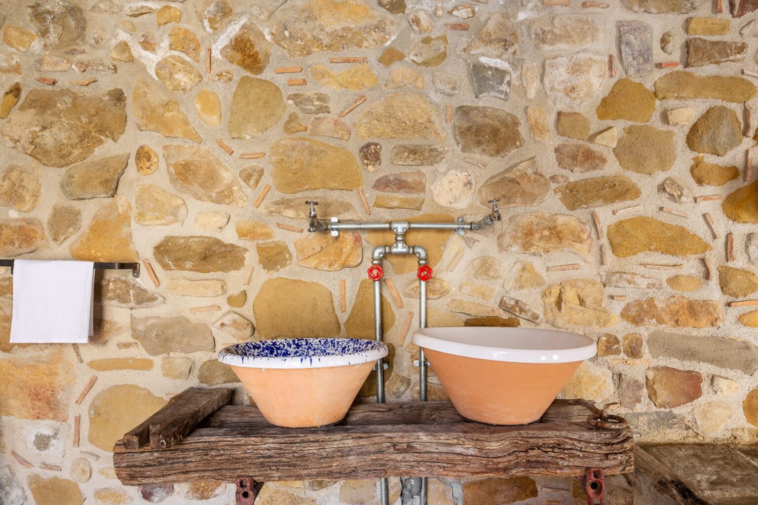 Rustic bathroom setup featuring two ceramic washbasins on a wooden countertop against a stone wall, with a vintage faucet and a white towel holder. | Only on SopranoVillas