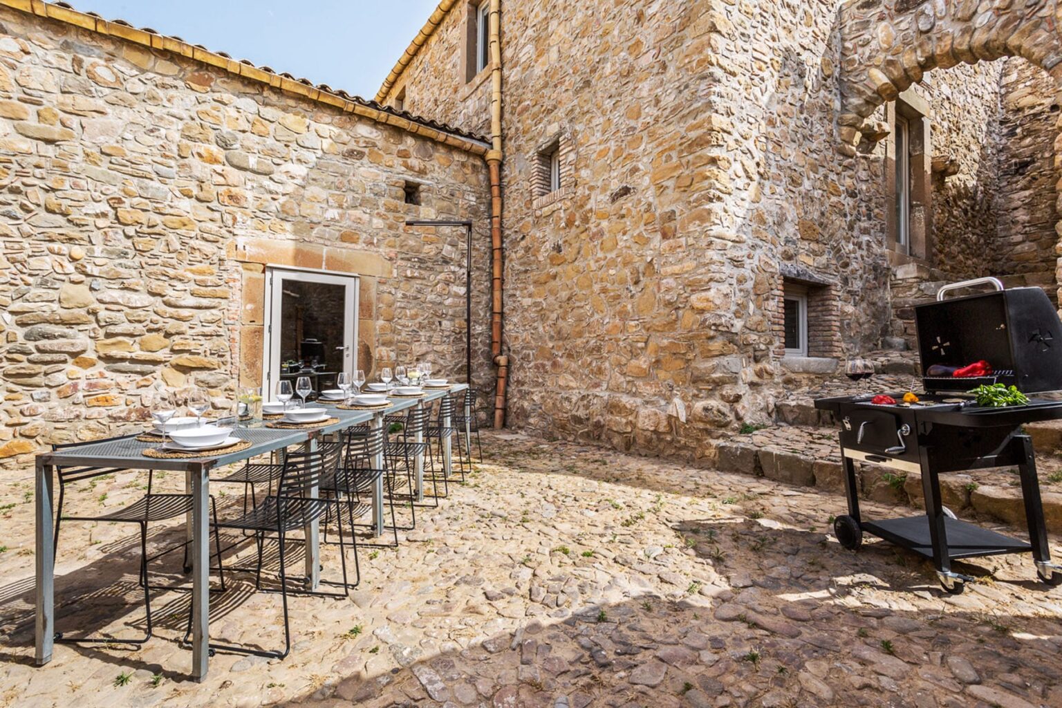 Outdoor dining area featuring a long table set with tableware next to a stone wall, with a barbecue grill in the foreground, ideal for gatherings and alfresco meals. | Only on SopranoVillas