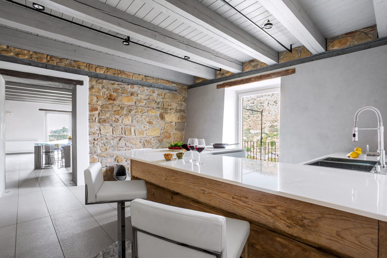 Modern kitchen with a sleek white countertop, wooden bar, and stone wall accents, featuring contemporary stools and large windows for natural light. | Only on SopranoVillas