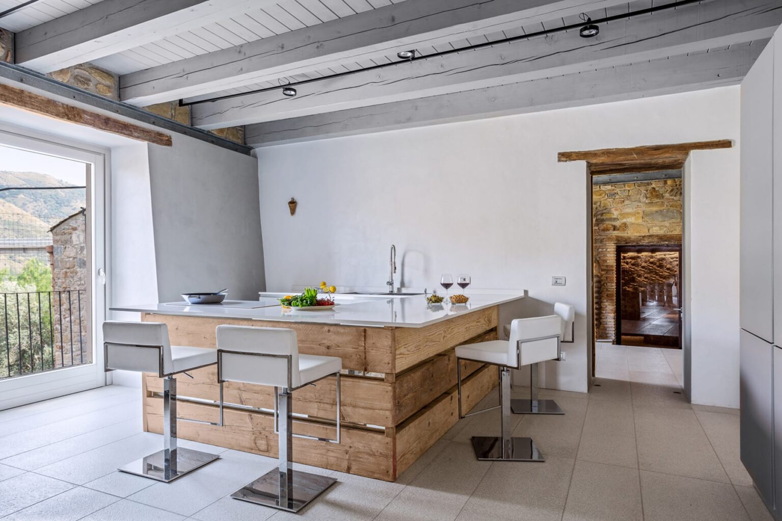 Modern kitchen featuring a wooden island, sleek white bar stools, and natural light from large windows. The design combines rustic elements with contemporary finishes, showcasing a minimalist aesthetic and open layout. | Only on SopranoVillas