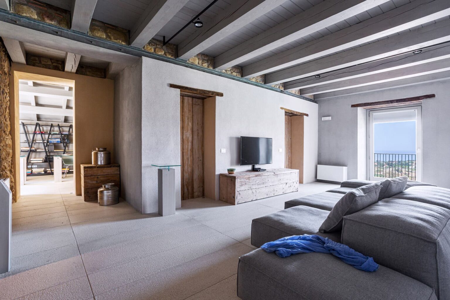 Modern living room with a minimalist design, featuring a large gray sofa, wooden doors, a flat-screen TV on a light wood media console, and a scenic view through a window. The interior showcases exposed beams and a neutral color palette, enhancing the spacious ambiance. | Only on SopranoVillas