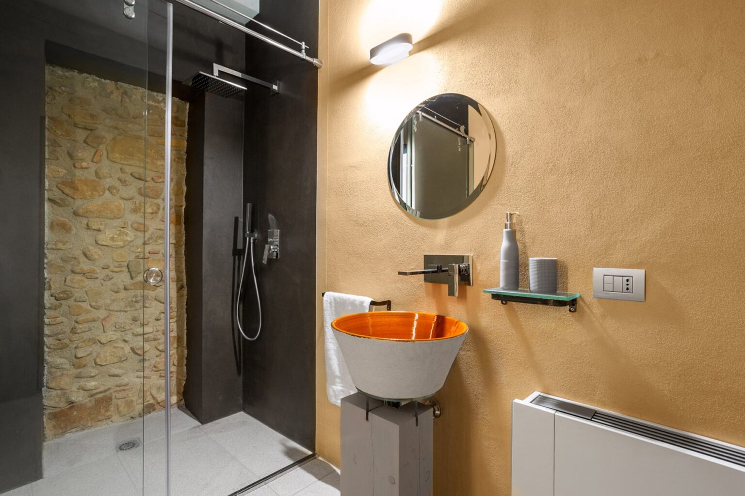 Modern bathroom design featuring a stylish orange sink, circular mirror, and sleek shower area with stone accents. The minimalist decor includes a glass shower door and contemporary fixtures, creating a bright and inviting space. | Only on SopranoVillas