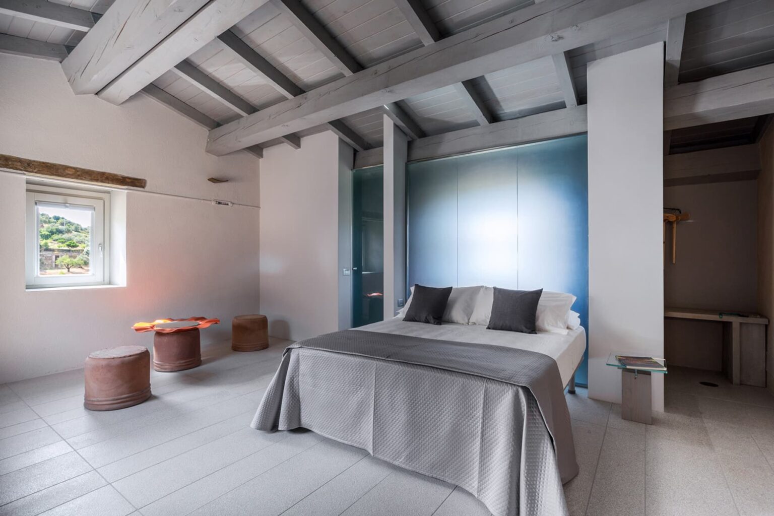 Modern bedroom interior featuring a comfortable bed with gray bedding, minimalistic decor, wooden beams, and stylish seating, creating a serene and inviting atmosphere. | Only on SopranoVillas