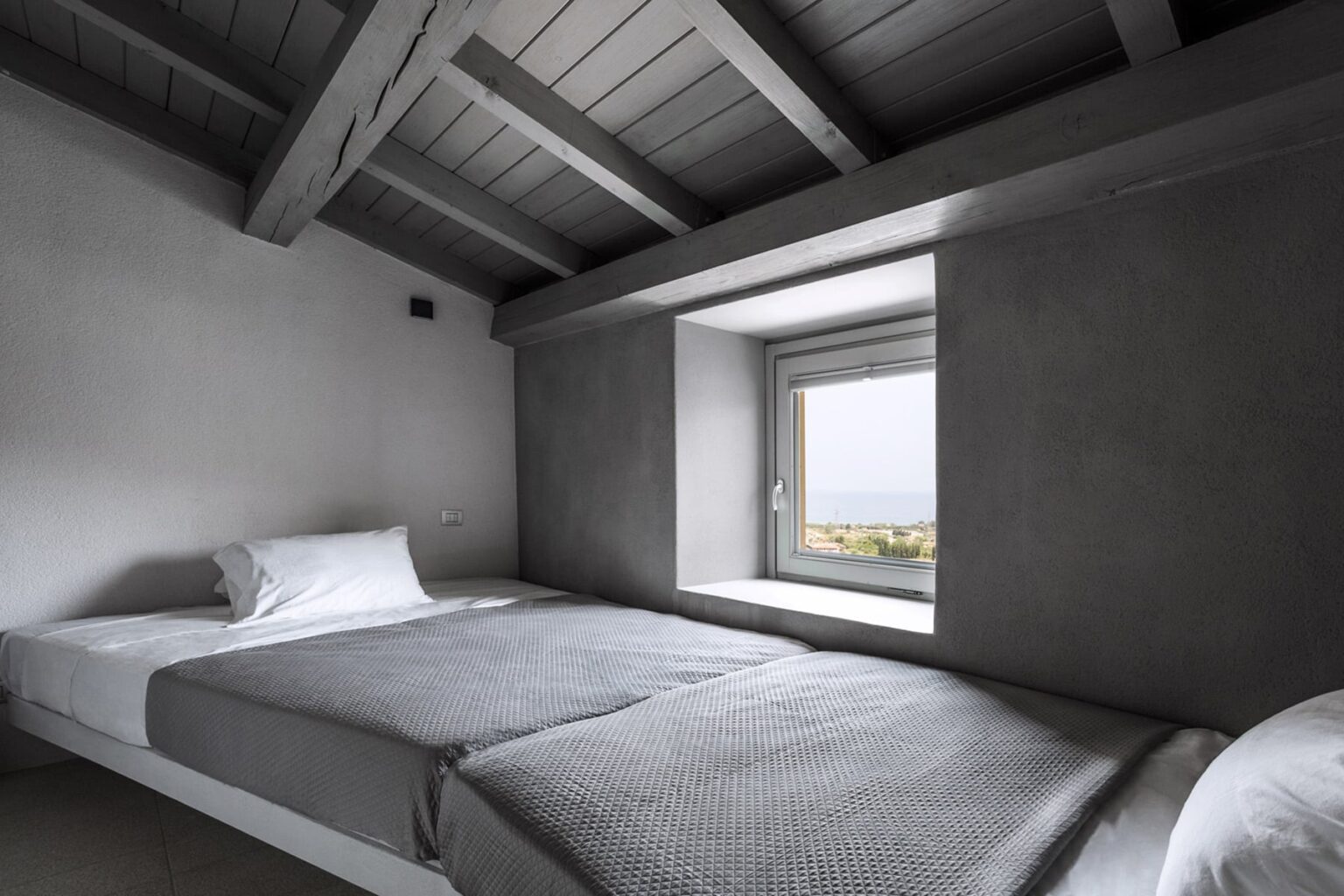 Minimalist bedroom with gray bedding, featuring a large window that offers a view of the outdoors, and exposed wooden beams on the ceiling. Ideal for modern interior design inspiration. | Only on SopranoVillas