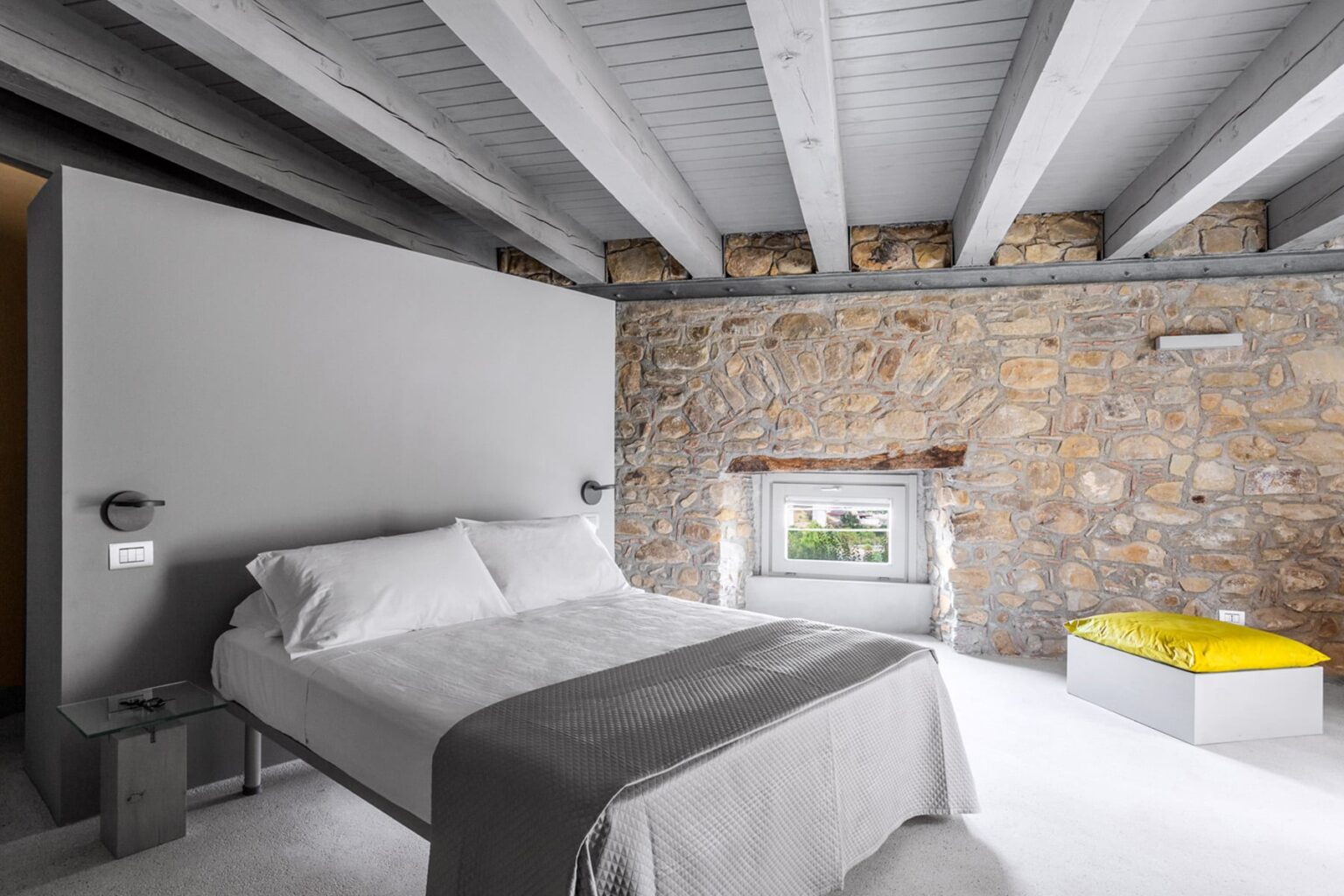 Modern bedroom design featuring a gray stone wall, minimalist furniture, a double bed with white linens, and a pop of color from a yellow accent piece. The room has exposed wooden beams and a window that enhances natural light. | Only on SopranoVillas