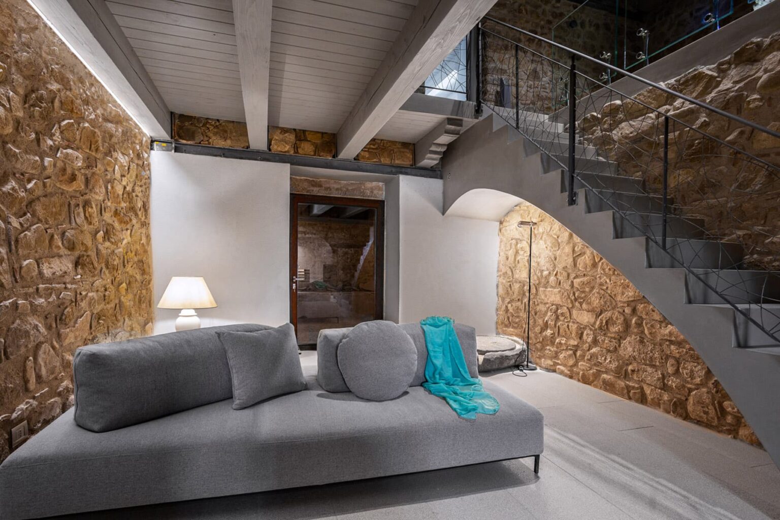 Modern living room featuring a gray sofa with decorative cushions, a turquoise throw, and a stylish lamp, set against rustic stone walls and a contemporary staircase. | Only on SopranoVillas