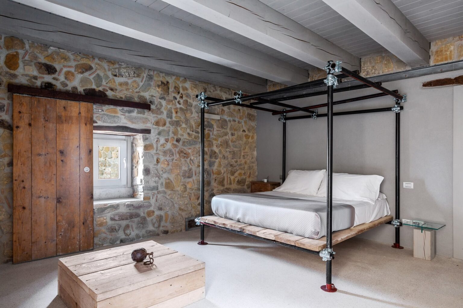 Stylish bedroom featuring a modern four-poster bed with a minimalist design, exposed wooden beams, and a rustic stone wall, complemented by natural light from a nearby window. | Only on SopranoVillas