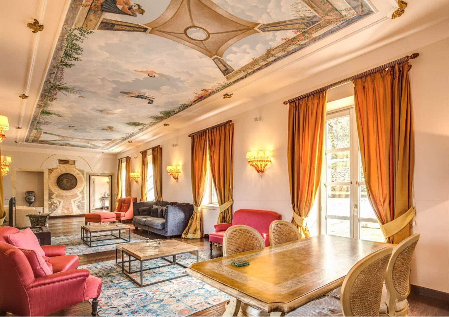 Elegant living room featuring a beautifully painted ceiling, warm orange curtains, and stylish seating. The space includes a wooden table and decorative rug, creating a cozy and inviting atmosphere. Ideal for relaxation and social gatherings. | Only on SopranoVillas
