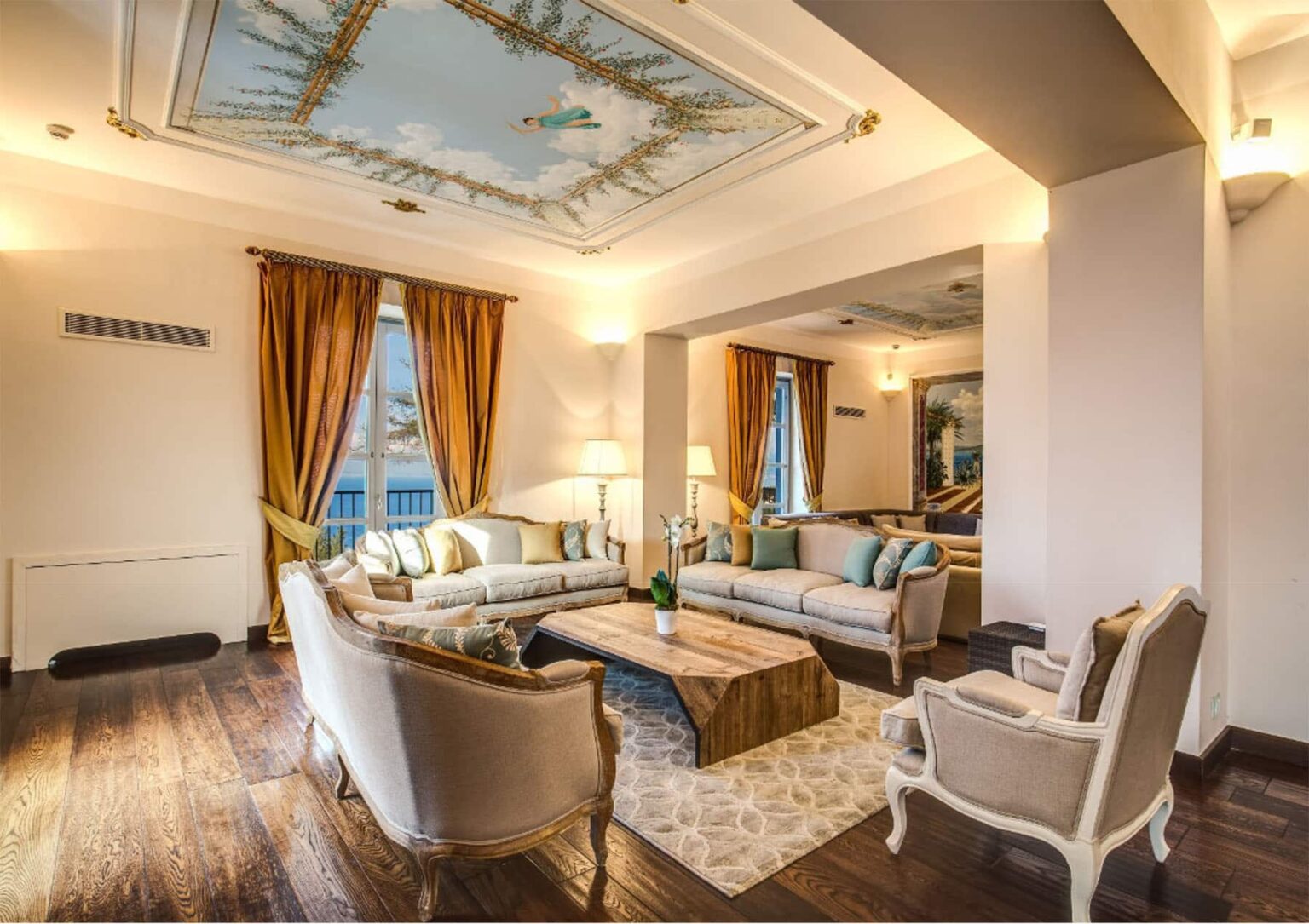 Elegant living room featuring plush sofas, a wooden coffee table, and decorative curtains. The ceiling showcases a beautiful mural, enhancing the room's ambiance with warm lighting and stylish decor. Ideal for relaxation and gatherings. | Only on SopranoVillas