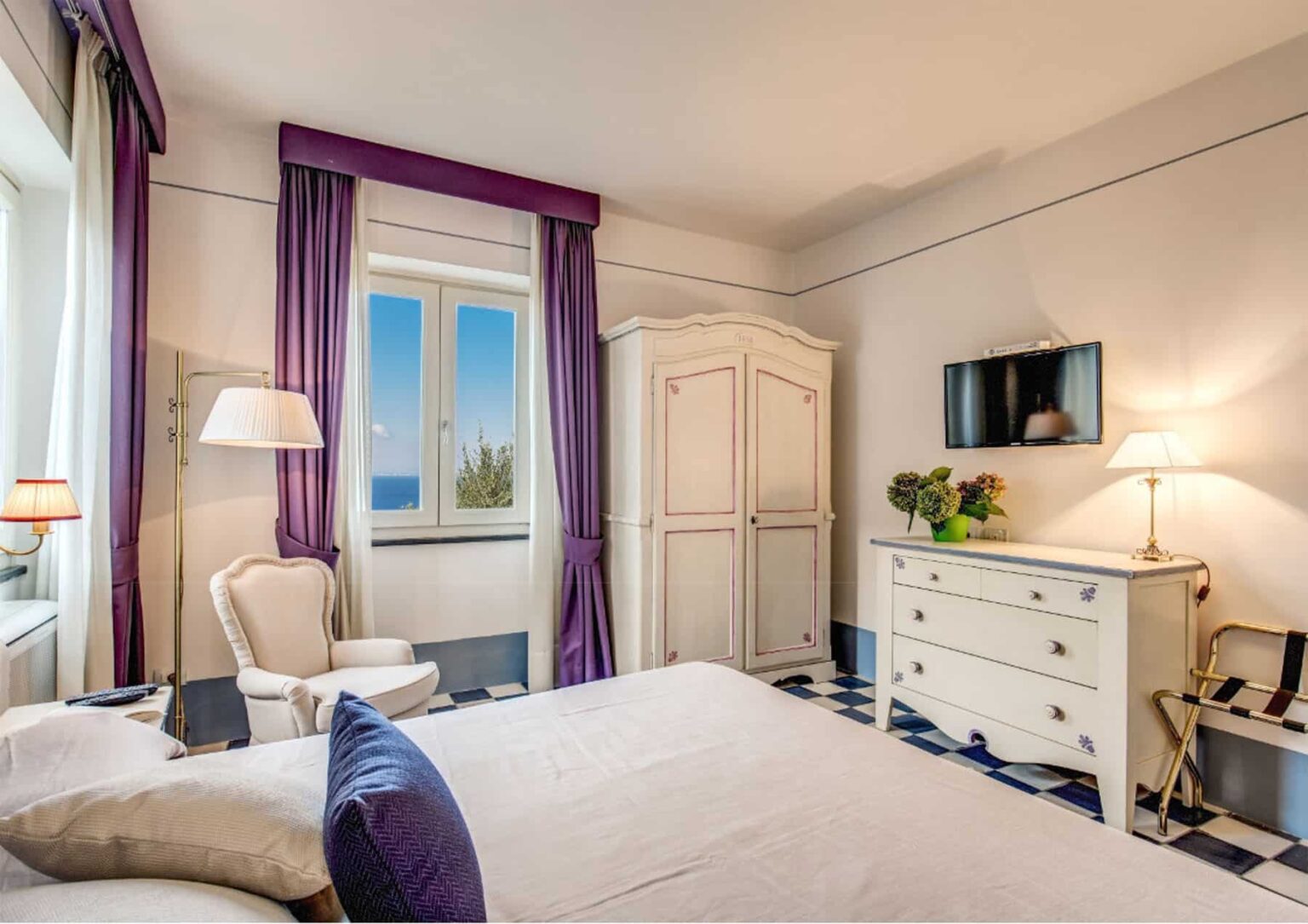 Cozy hotel room featuring a queen-sized bed, elegant white furniture, and large windows with scenic views. The room is decorated with purple accents, a comfortable armchair, and a flat-screen TV, creating a relaxing atmosphere for guests. | Only on SopranoVillas