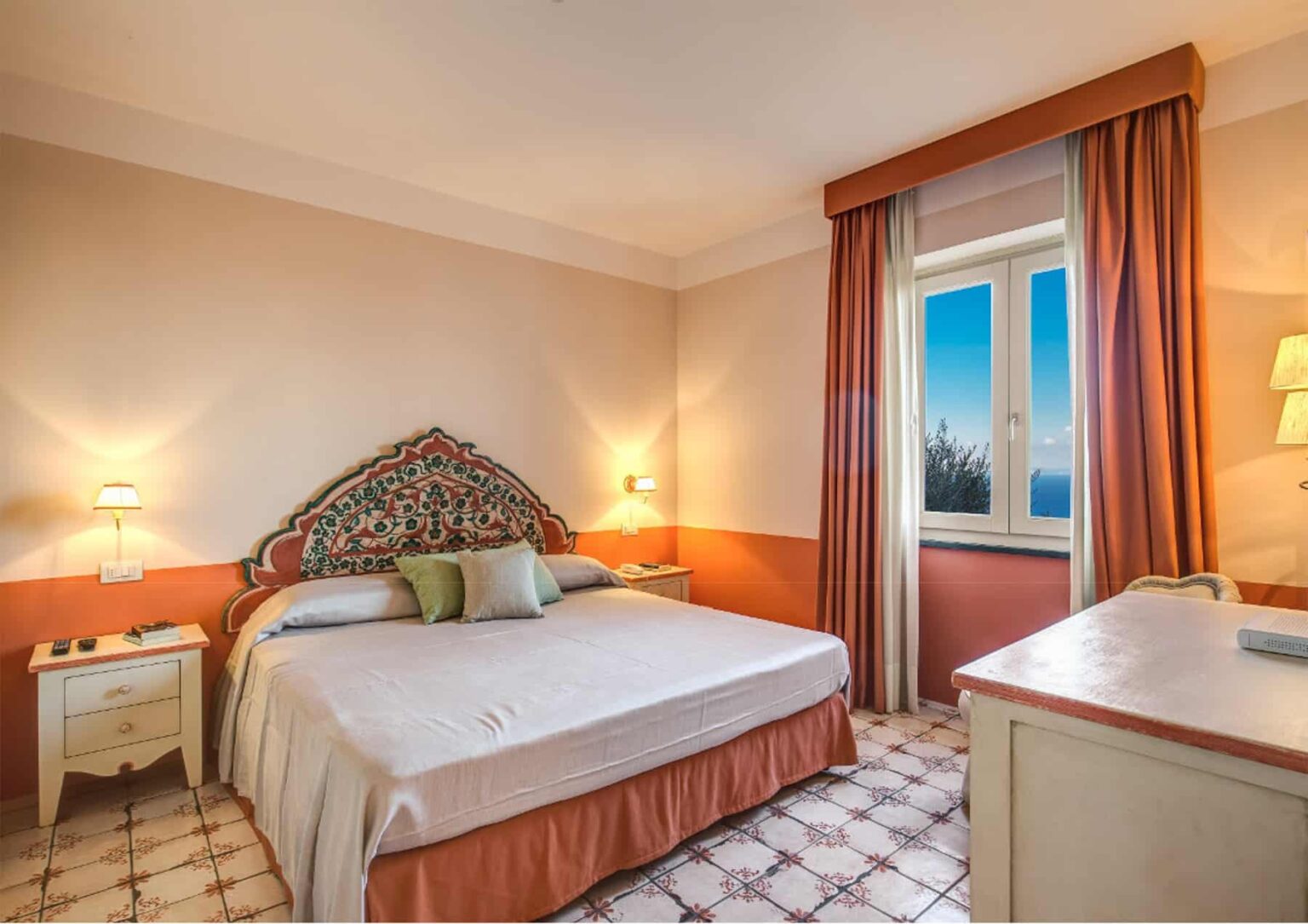 Cozy hotel bedroom featuring a large bed with decorative headboard, warm lighting, and a window with scenic views. The room is adorned with soft colors and elegant furnishings, creating a welcoming atmosphere for guests. | Only on SopranoVillas
