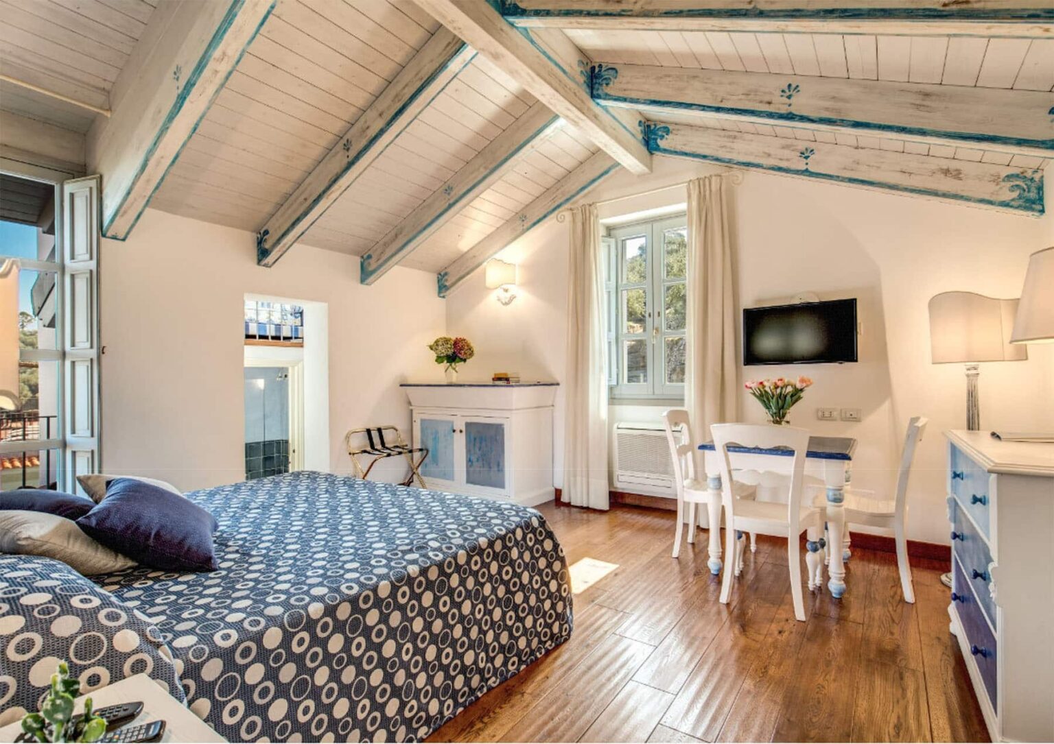 Cozy bedroom with high wooden ceilings, a patterned bedspread, a small dining area, and a flat-screen TV, featuring large windows that allow natural light. Ideal for a relaxing getaway. | Only on SopranoVillas