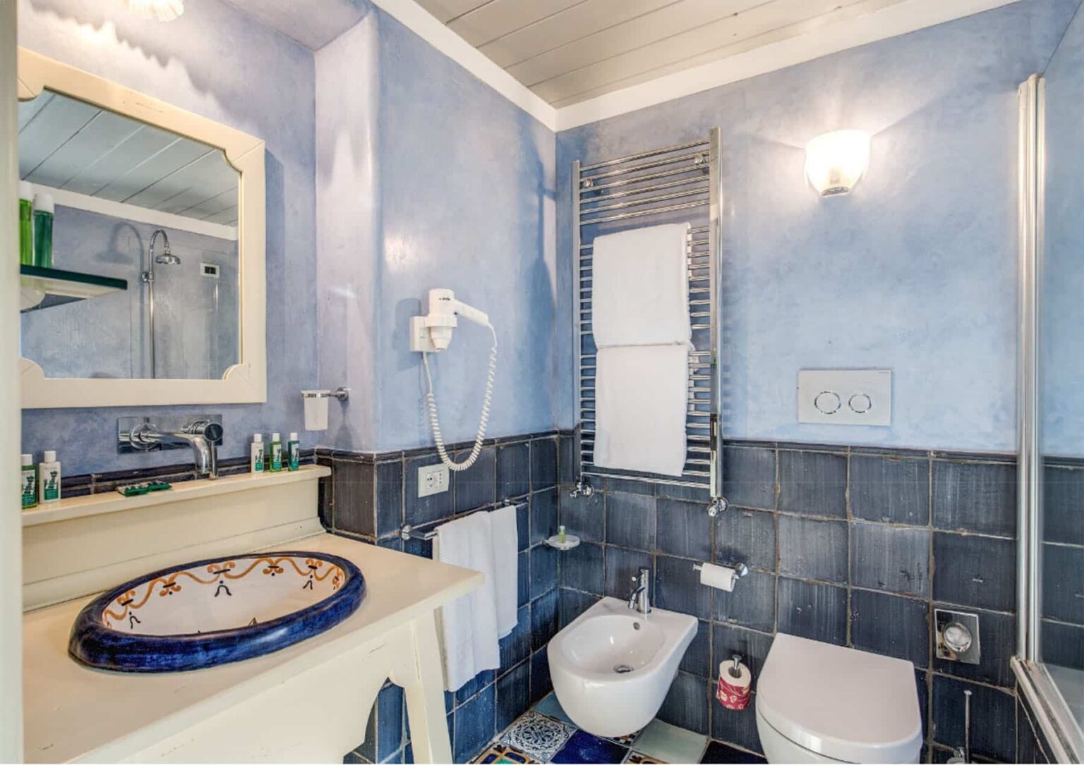 Modern bathroom featuring blue walls, a stylish sink with decorative edges, a bidet, and a glass shower enclosure, complemented by a mirror and various toiletries. | Only on SopranoVillas