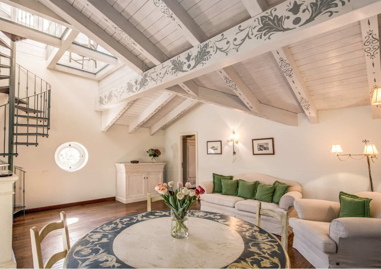 Spacious living room with high vaulted ceiling, decorative wooden beams, and a spiral staircase. The room features a round table with a floral centerpiece, a comfortable sofa adorned with green cushions, and soft lighting, creating a cozy and inviting atmosphere. | Only on SopranoVillas