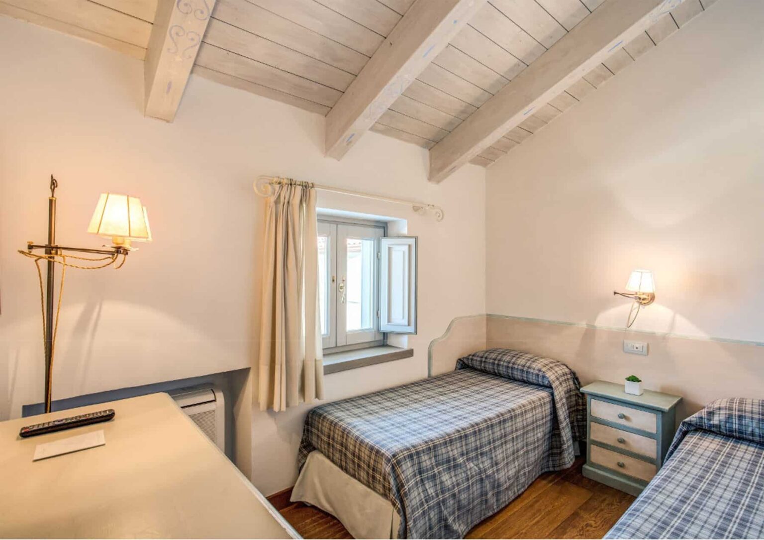 Cozy bedroom with two twin beds, wooden beams, a desk, and natural light from a window, featuring light-colored walls and a bedside lamp. | Only on SopranoVillas