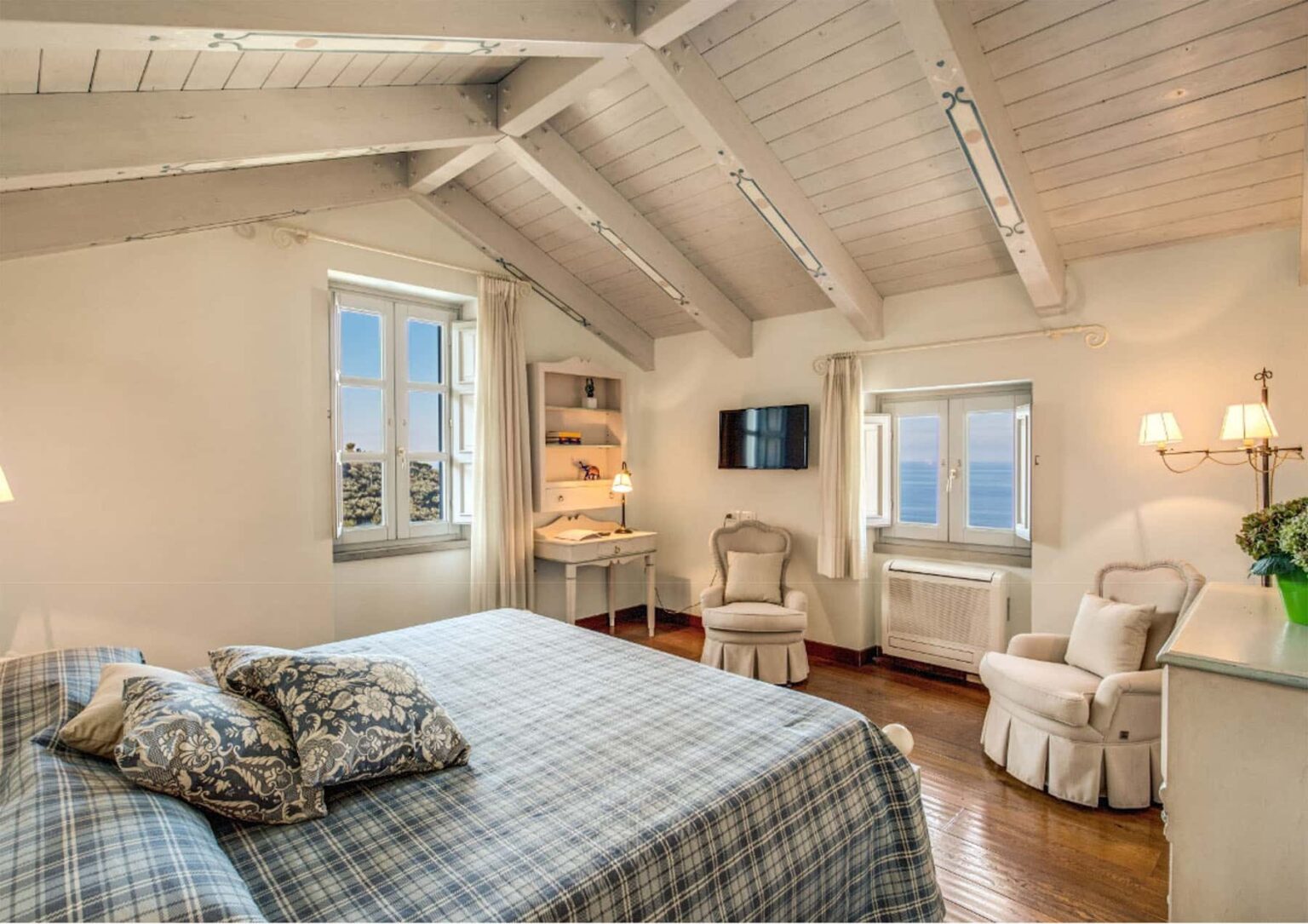 Cozy bedroom with a large bed, stylish decor, and a view of the sea through two windows, featuring wooden beams and modern amenities like a TV and air conditioning. | Only on SopranoVillas