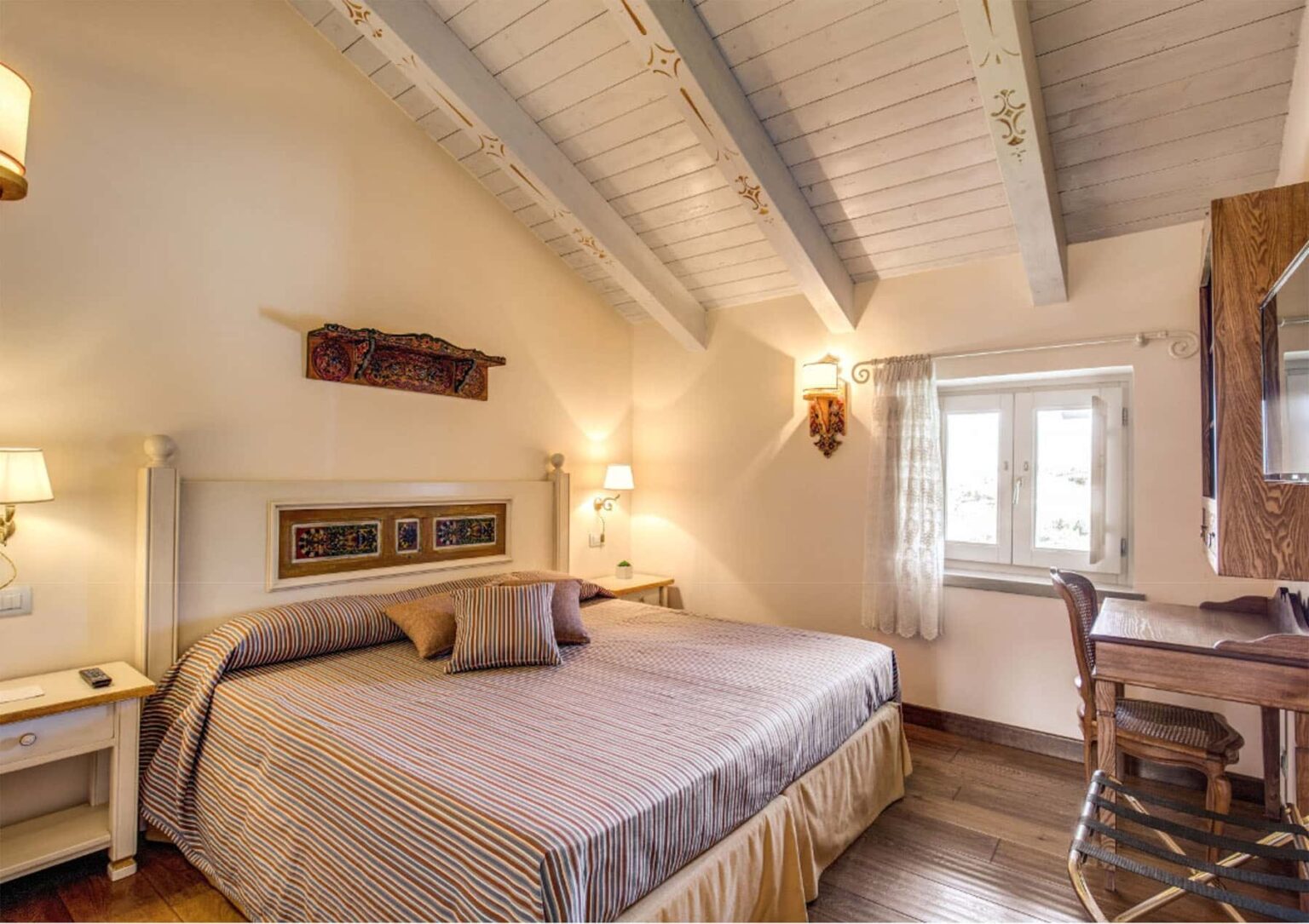 Cozy hotel room featuring a queen-sized bed with decorative pillows, wooden beams, and a vintage desk. Natural light streams in through the window, enhancing the warm, inviting atmosphere. Ideal for travelers seeking comfort and charm. | Only on SopranoVillas