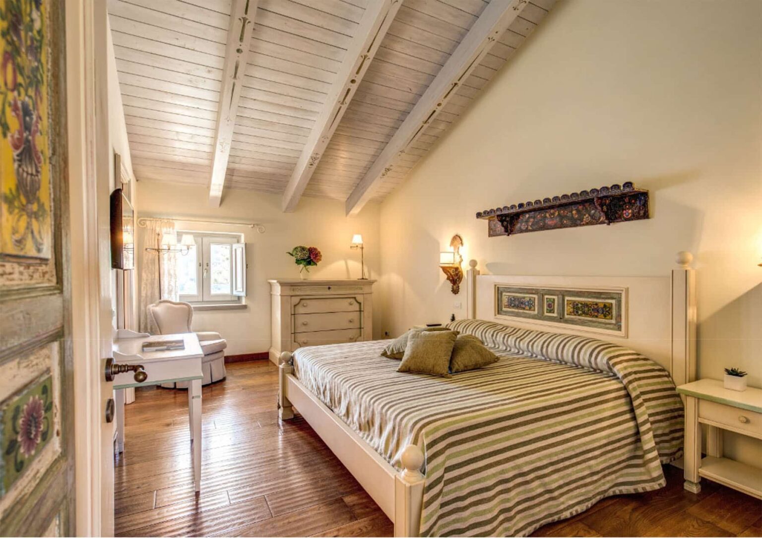 Cozy bedroom with a striped bedspread, wooden floors, and exposed beam ceiling, featuring a dresser, bedside lamps, and natural light from large windows. | Only on SopranoVillas