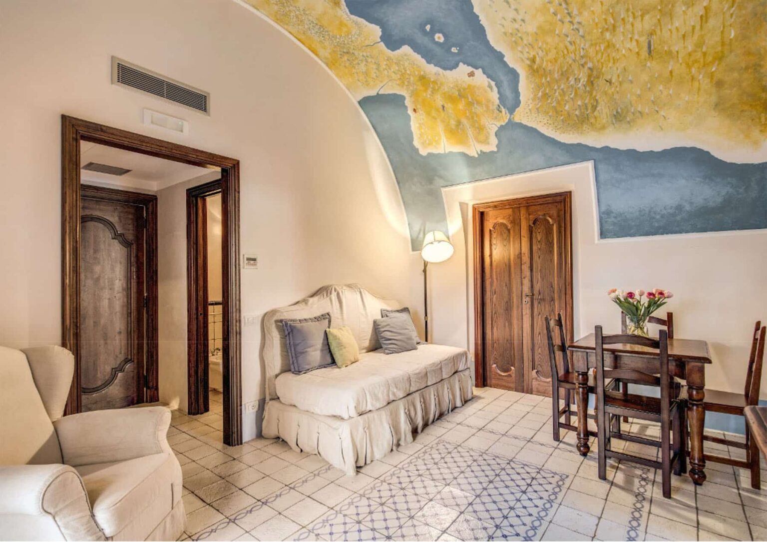 Cozy living area featuring a mural of a map on the ceiling, a comfortable sofa with decorative pillows, a wooden dining table, and fresh flowers in a vase, designed for relaxation and style. | Only on SopranoVillas