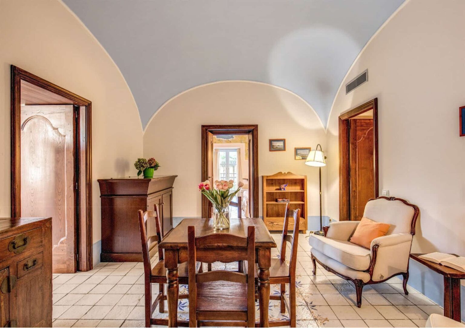 Cozy dining area featuring a wooden table with chairs, a sideboard, and a comfortable armchair, illuminated by natural light from a nearby window, showcasing an inviting and warm interior design. | Only on SopranoVillas