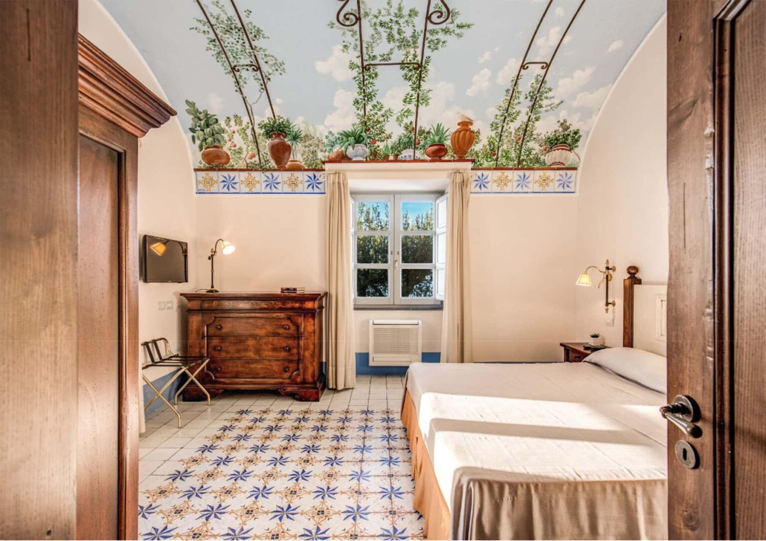 Cozy bedroom with a decorative ceiling featuring plants, a large window, and traditional furnishings including a wooden dresser and patterned floor tiles. Ideal for showcasing interior design and home decor. | Only on SopranoVillas