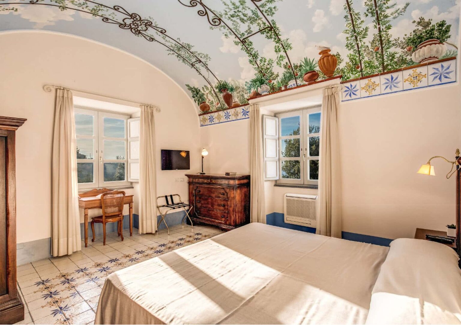 Cozy hotel room featuring a double bed, elegant wooden furniture, large windows with scenic views, and a beautifully painted ceiling adorned with greenery and floral designs. | Only on SopranoVillas
