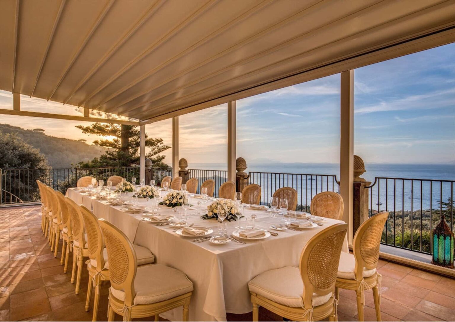 Elegant dining setup with a long table adorned with white tablecloth and floral centerpieces, overlooking a scenic coastal view through large glass windows during sunset. | Only on SopranoVillas
