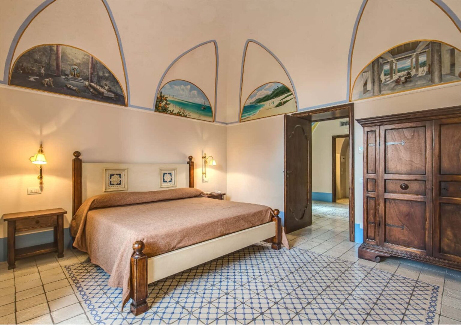 Cozy bedroom featuring a double bed with brown linens, traditional wooden furniture, and decorative arched wall paintings. The room is well-lit with soft lighting and has a charming tiled floor, creating a warm and inviting atmosphere. | Only on SopranoVillas