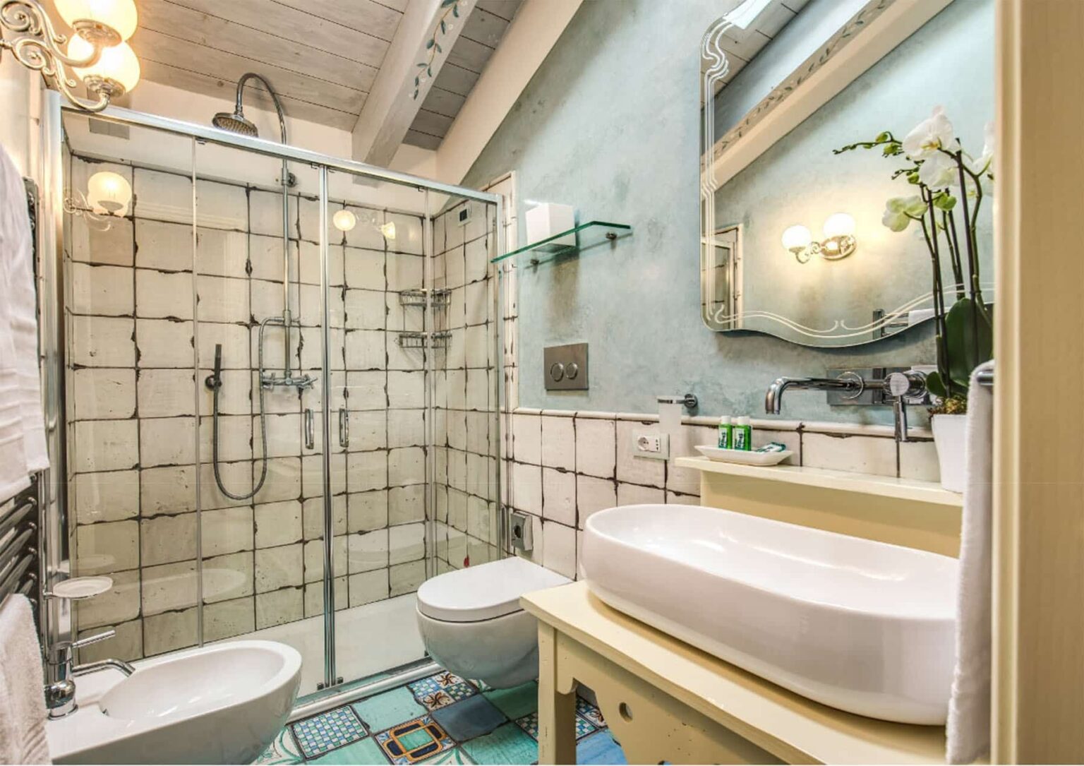 Modern bathroom featuring a glass shower enclosure, elegant sink with countertop, stylish mirror, and decorative plants, all set against a light blue wall and tiled accents. | Only on SopranoVillas