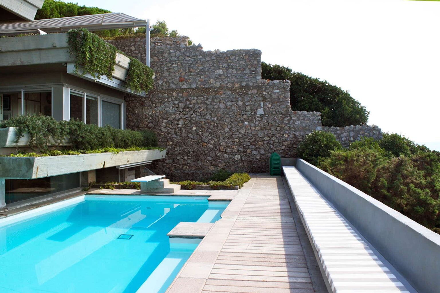 Modern luxury villa with a sleek swimming pool and stone wall landscaping, surrounded by greenery and featuring a wooden deck. Ideal for outdoor relaxation and entertaining. | Only on SopranoVillas