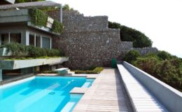 Modern luxury villa with a sleek swimming pool and stone wall landscaping, surrounded by greenery and featuring a wooden deck. Ideal for outdoor relaxation and entertaining.