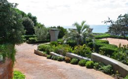Scenic pathway leading through lush greenery and vibrant flowers, with a view of the ocean in the background, showcasing a tranquil outdoor setting.