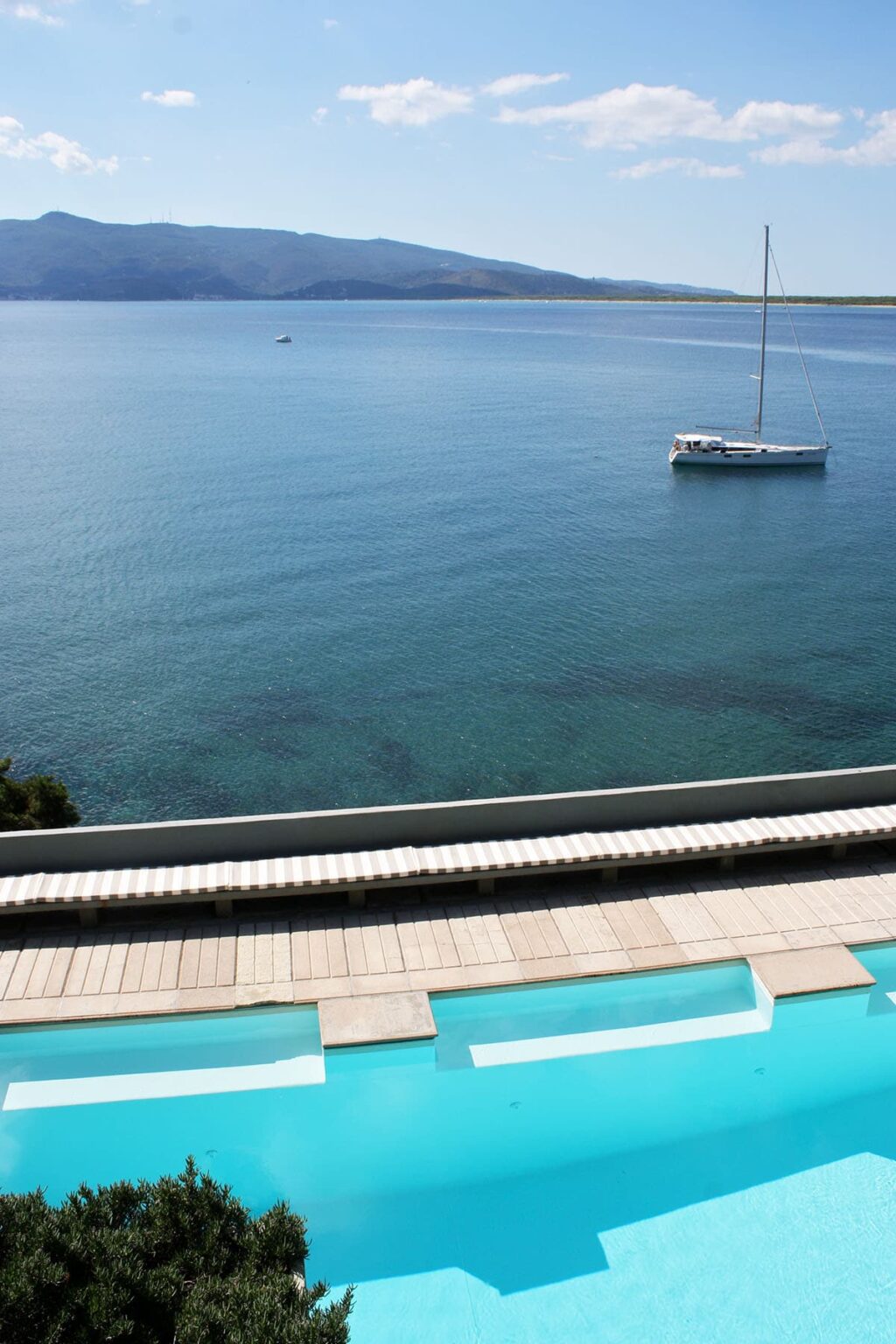 Scenic view of calm blue waters with a white boat anchored offshore and a sunlit shoreline featuring lounge chairs. | Only on SopranoVillas