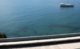 Scenic view of calm blue waters with a white boat anchored offshore and a sunlit shoreline featuring lounge chairs.