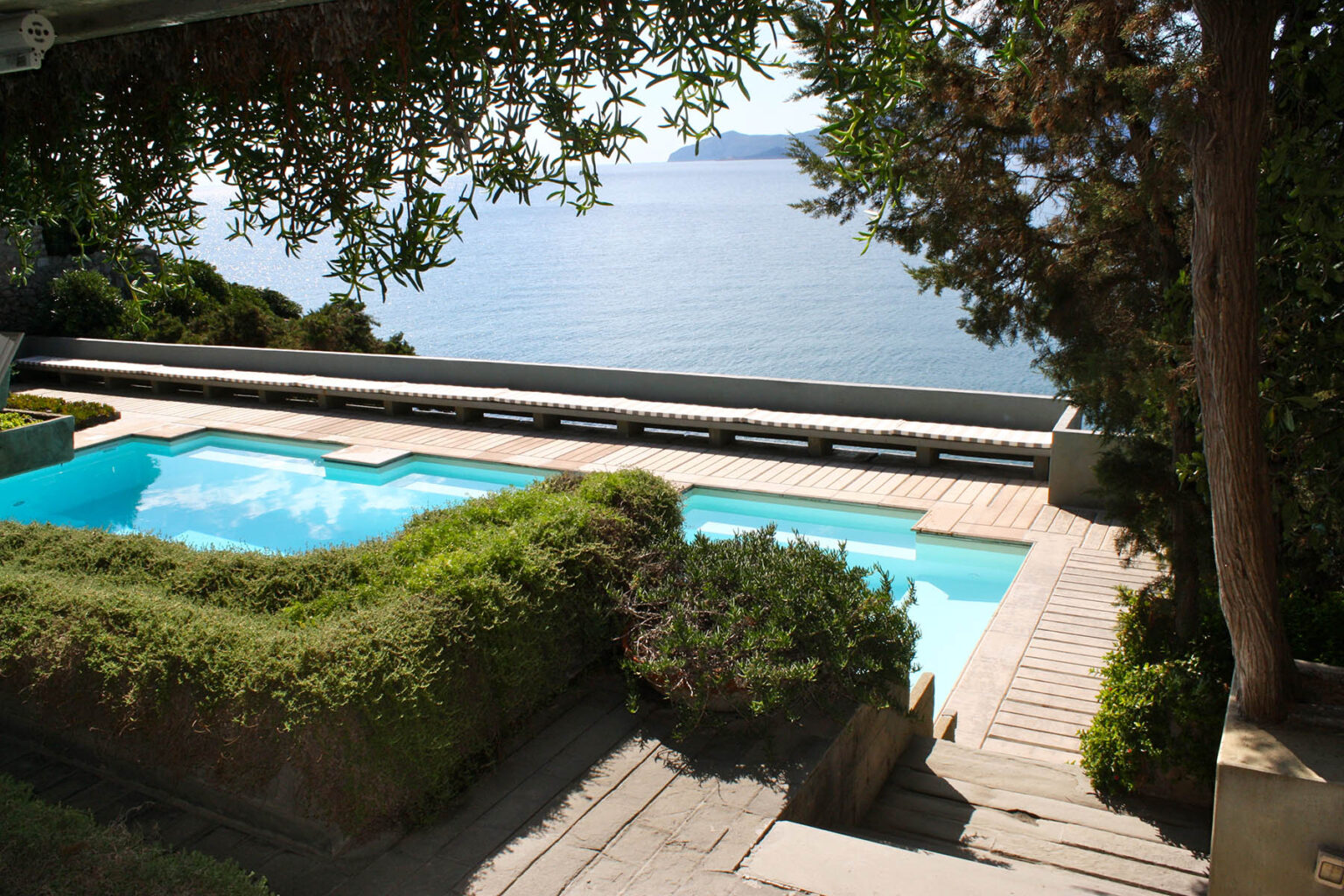 Scenic view of a tranquil poolside area overlooking a serene body of water, surrounded by lush greenery and stone pathways, perfect for relaxation and outdoor enjoyment. | Only on SopranoVillas