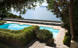 Scenic view of a tranquil poolside area overlooking a serene body of water, surrounded by lush greenery and stone pathways, perfect for relaxation and outdoor enjoyment.