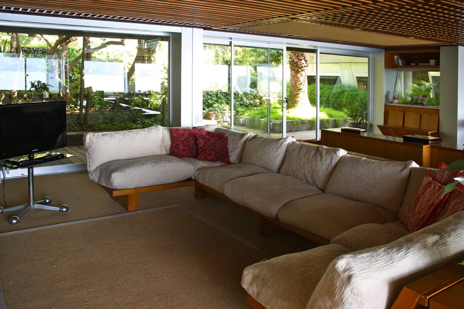 Modern living room with large windows overlooking a garden, featuring a spacious beige sectional sofa with colorful cushions and a sleek television stand. Bright natural light fills the space, creating a warm and inviting atmosphere. | Only on SopranoVillas