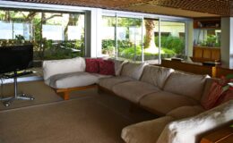 Modern living room with large windows overlooking a garden, featuring a spacious beige sectional sofa with colorful cushions and a sleek television stand. Bright natural light fills the space, creating a warm and inviting atmosphere.