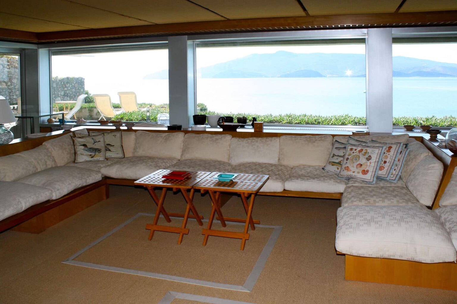 Cozy living room with a large sectional sofa, a wooden coffee table, and panoramic windows offering a view of the ocean and mountains. Natural light floods the space, creating a warm and inviting atmosphere. | Only on SopranoVillas