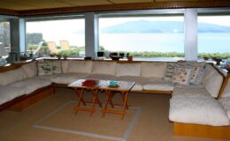 Cozy living room with a large sectional sofa, a wooden coffee table, and panoramic windows offering a view of the ocean and mountains. Natural light floods the space, creating a warm and inviting atmosphere.
