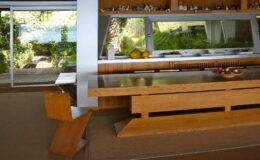 Modern kitchen interior featuring a sleek wooden dining table, minimalist design, and large windows that provide natural light and views of greenery outside.